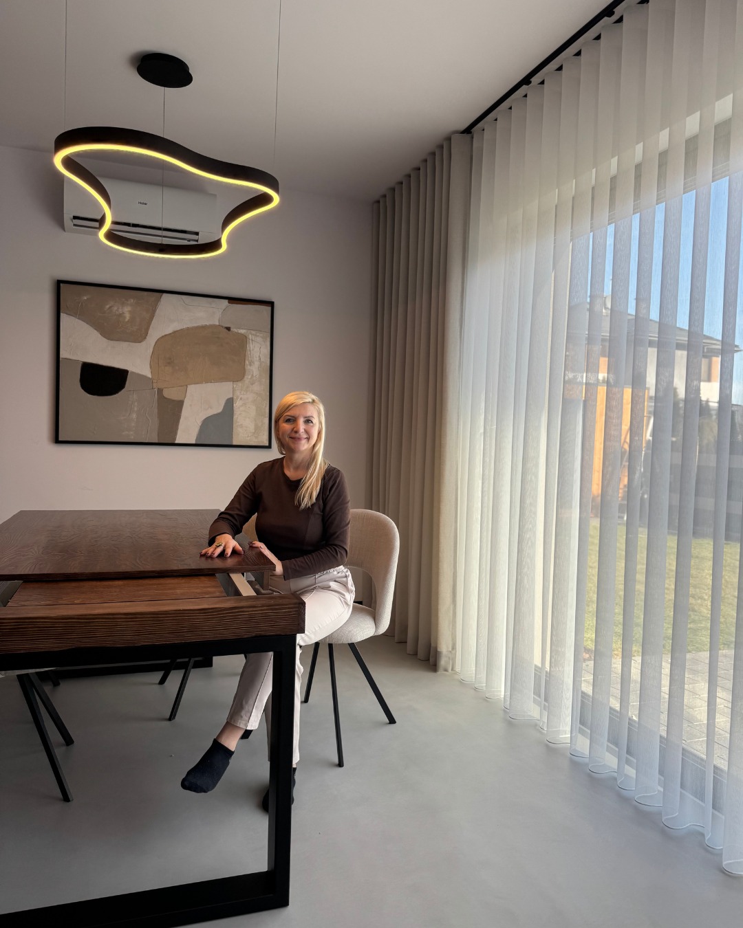 Kobieta siedzi przy drewnianym stole na tle okna z pionowymi, jasnymi żaluzjami panelowymi, nad stołem nowoczesna lampa z żółtym światłem.