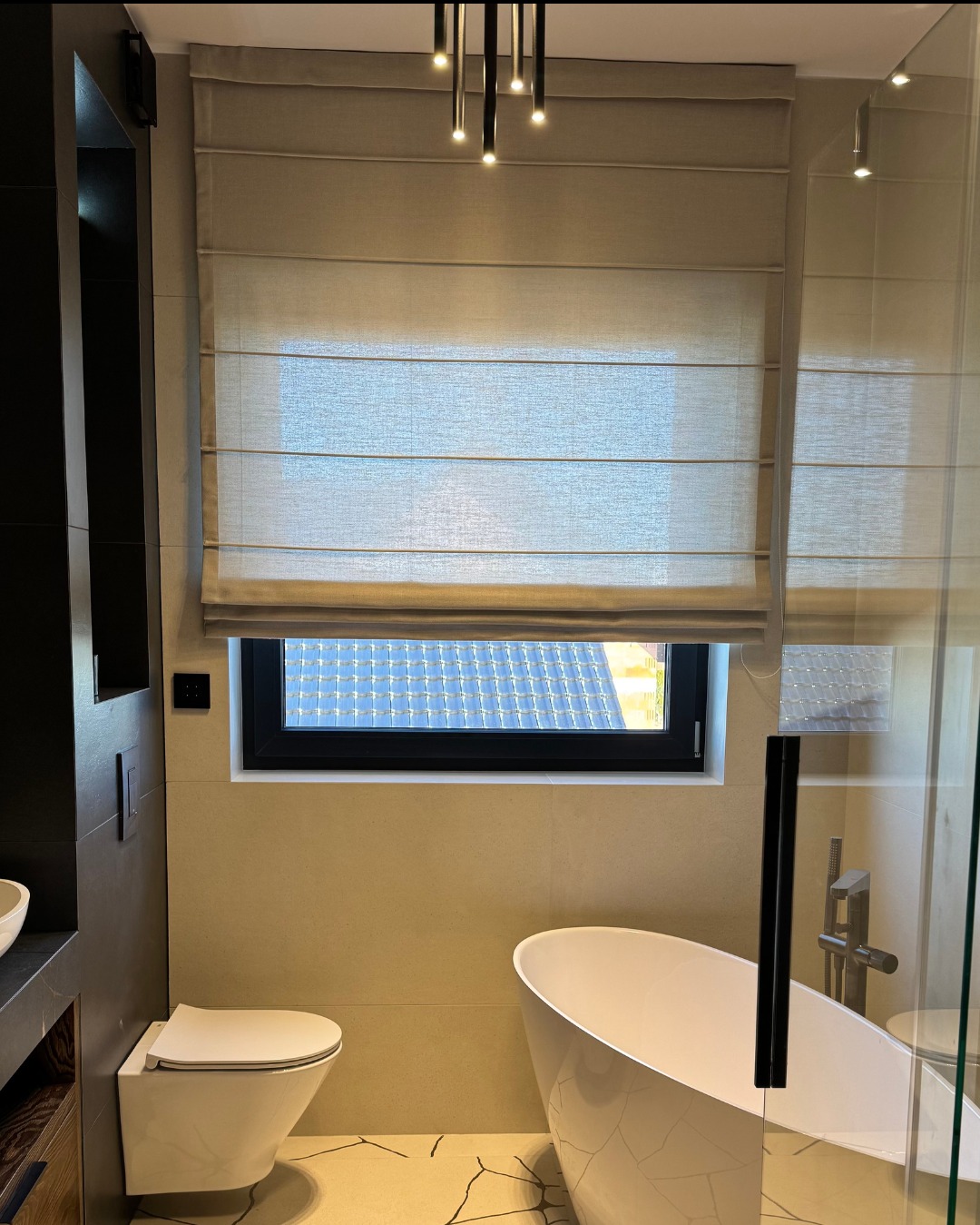 Beżowa, płaska roleta w minimalistycznej łazience z wanną wolnostojącą i widokiem na dach za oknem.