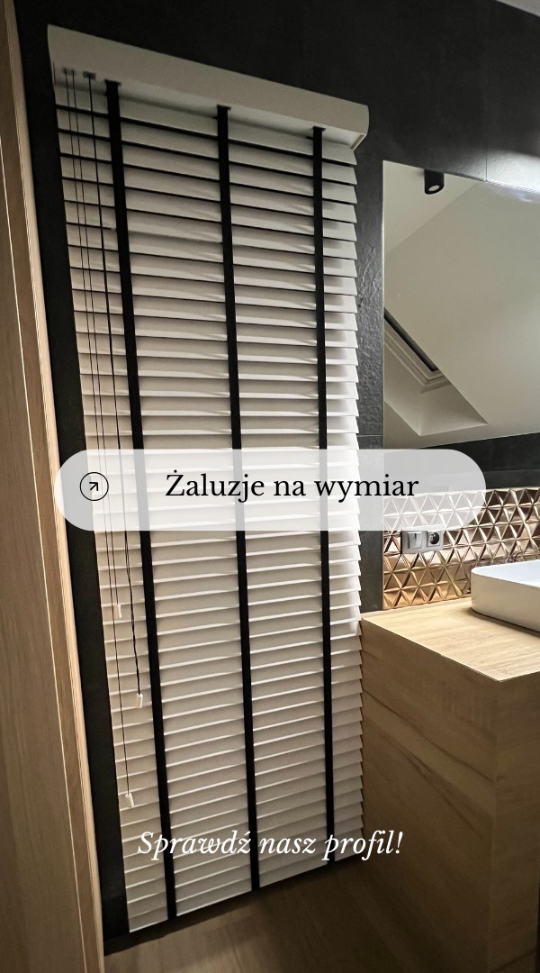 Białe żaluzje drewniane z czarnymi tasiemkami zamontowane w nowoczesnej łazience z elementami dekoracyjnymi w kolorze złota.