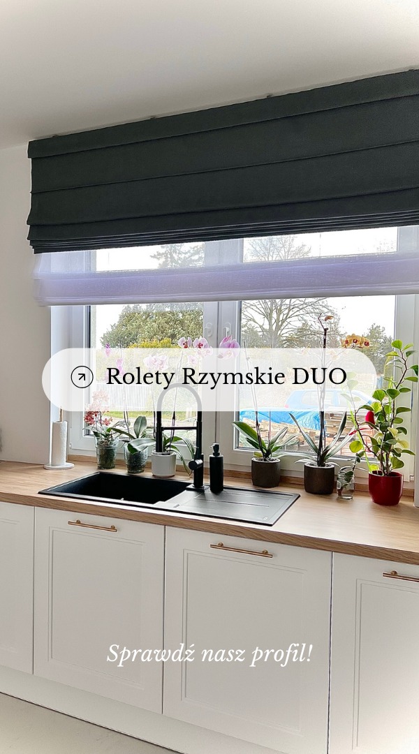 Ciemnoszara roleta rzymska typu dzień-noc w kuchni z białymi szafkami, drewnianym blatem i kwiatami w doniczkach na parapecie.