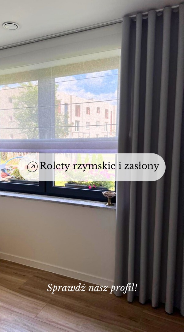 Szara, gruba zasłona z regularnymi fałdami po prawej stronie okna, obok roleta rzymska w neutralnym odcieniu, przez którą widać budynek i drzewa.