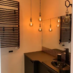 Czarna szafka łazienkowa z umywalką wpuszczaną w blat, podświetlona wiszącymi lampami z żarówkami Edisona, obok grzejnik drabinkowy.