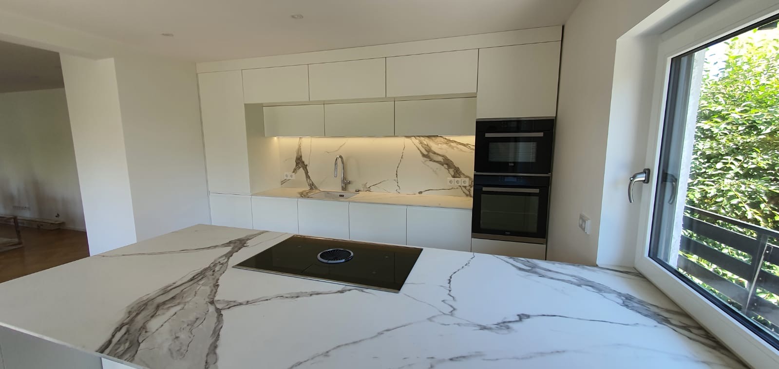 Nowoczesna biała kuchnia z marmurowym blatem wyspowym i backsplashiem, zintegrowany piekarnik i mikrofalówka, widok na zieleń za oknem.