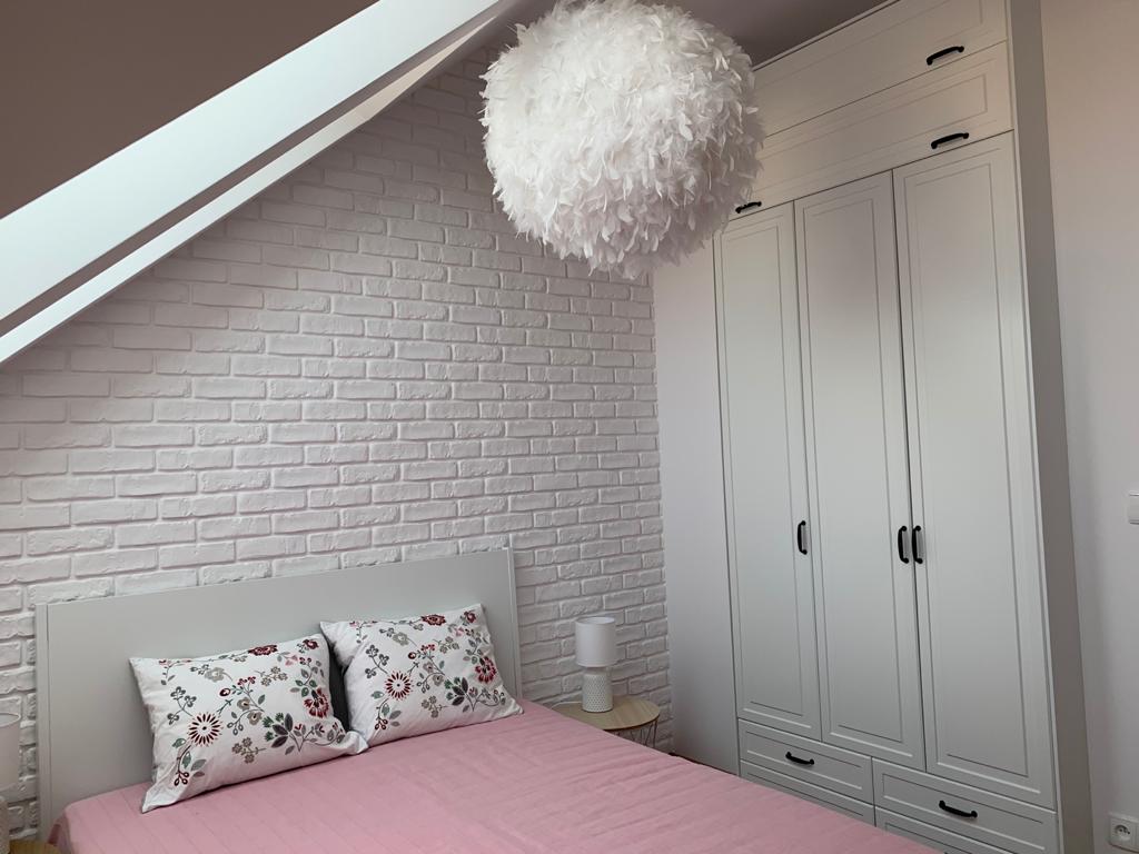 Biała szafa wnękowa z trzema drzwiami i szufladami w sypialni na poddaszu z ceglaną ścianą i dekoracyjną lampą z piór.