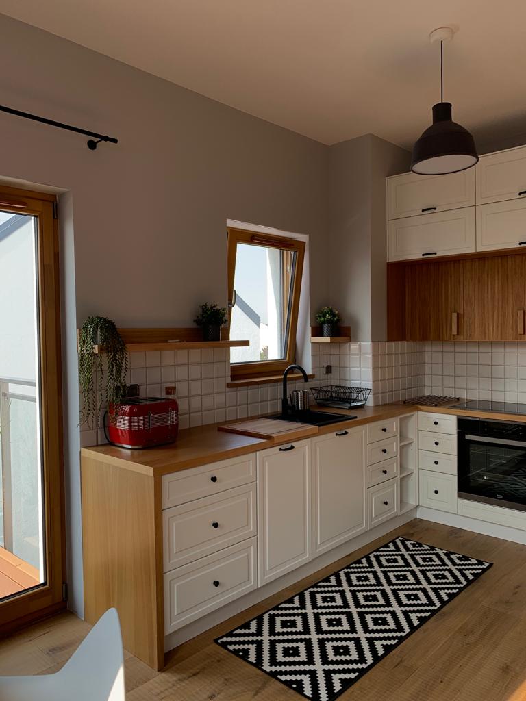 Jasna kuchnia z białymi szafkami z drewnianymi blatami, czarną armaturą i geometrycznym dywanem na podłodze.