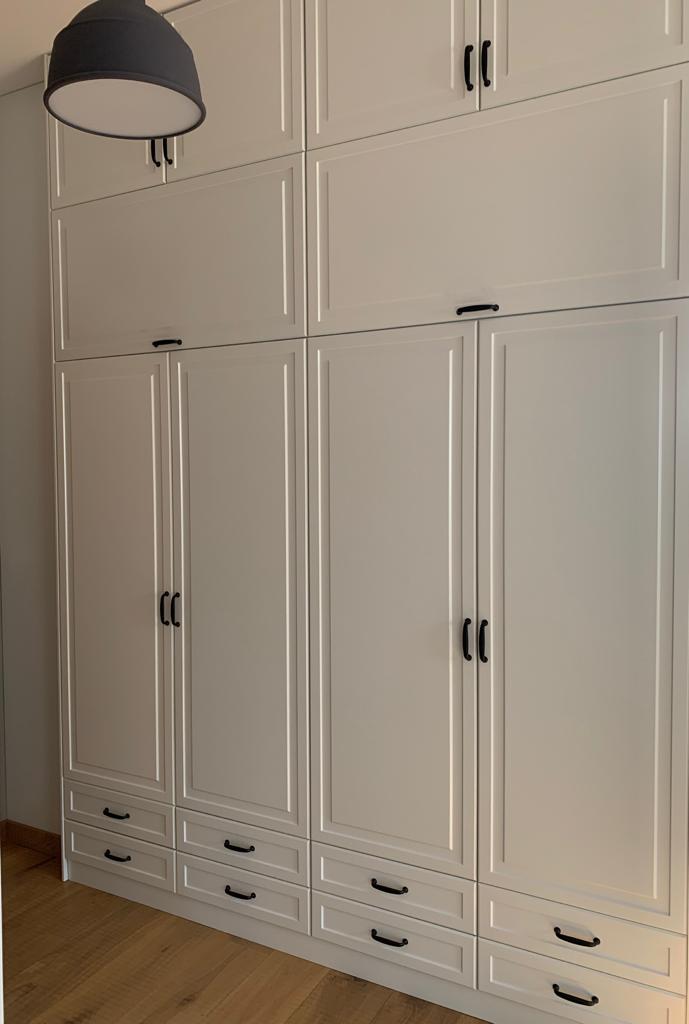 Duża, biała szafa wnękowa z czarnymi uchwytami, podzielona na segmenty z szafkami i szufladami, oświetlona przez wiszącą lampę z szarym abażurem.