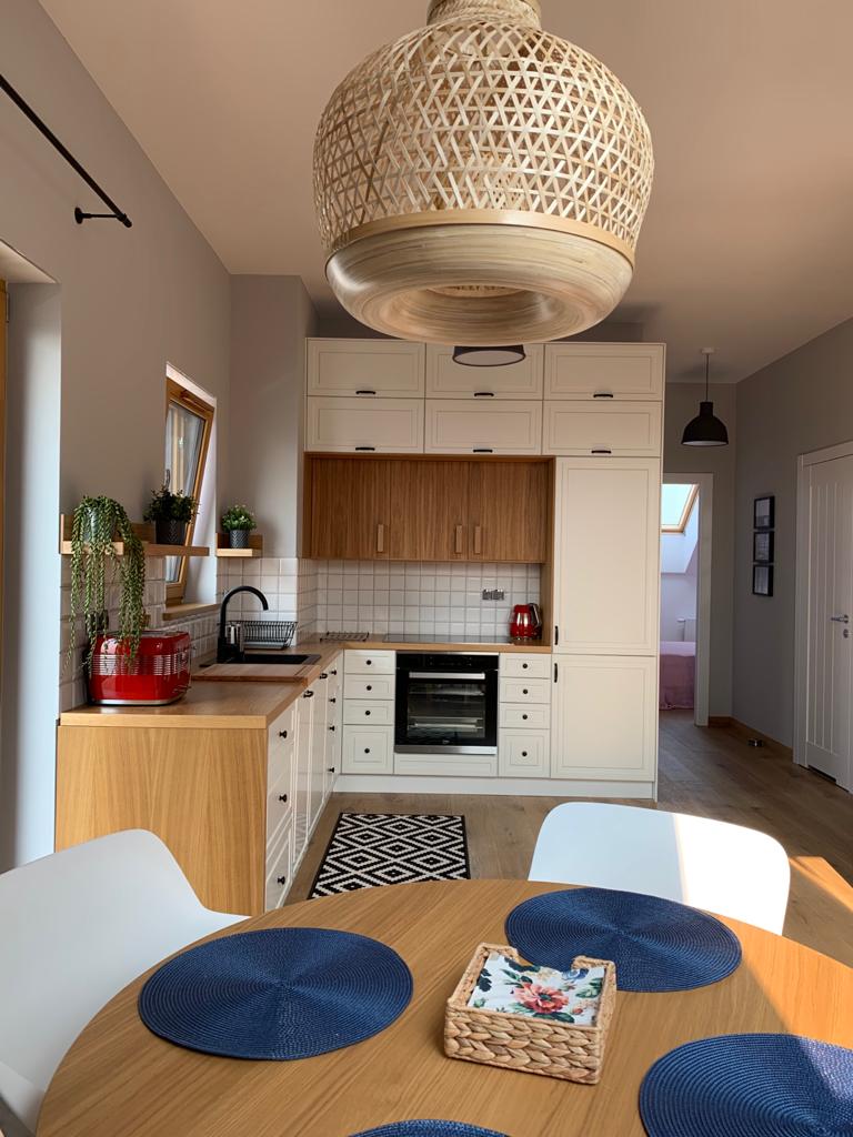 Jasna kuchnia z drewnianymi akcentami w Wysogotowie: zabudowa kuchenna z białymi frontami i drewnianymi szafkami, okrągły stół z niebieskimi podkładkami, pleciona lampa wisząca.