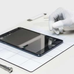 Naprawa Telefonów Tabletów Lublin - Uszkodzony tablet leży na białej macie z siatką, obok precyzyjny śrubokręt i narzędzie do otwierania obudów, dłoń w rękawiczce antystatycznej z niebieską opaską na nadgarstku dotyka ekranu.