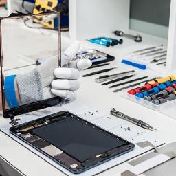 Naprawa Telefonów Tabletów Lublin - Technik w rękawicy trzyma uszkodzony ekran tabletu nad rozłożonym urządzeniem; widoczne narzędzia i śrubki na białym stole.