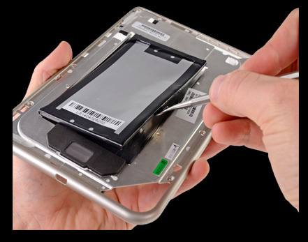 Otwarta obudowa tabletu z wyjmowaną baterią, podważaną metalowym narzędziem przez dłoń technika.