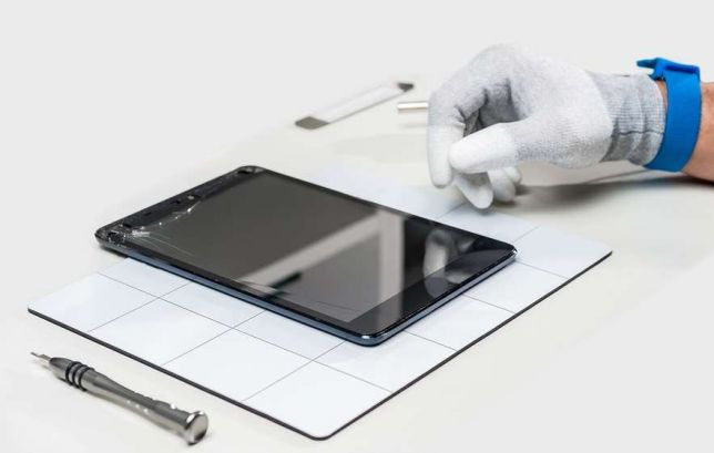 Uszkodzony tablet leży na białej macie z siatką, obok precyzyjny śrubokręt i narzędzie do otwierania obudów, dłoń w rękawiczce antystatycznej z niebieską opaską na nadgarstku dotyka ekranu.