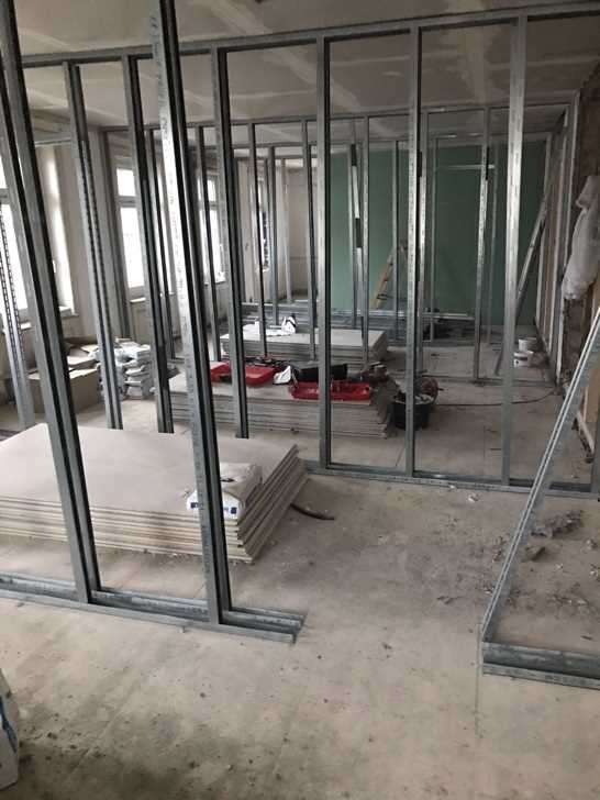 Wnętrze w trakcie remontu: widoczne metalowe profile konstrukcyjne tworzące szkielet ścian działowych i stos płyt na podłodze, przygotowanych do montażu.