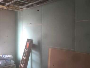 Wnętrze w trakcie remontu: ściany wyłożone płytami, widoczna drabina oparta o ścianę, sufit z metalową konstrukcją, worki z materiałami budowlanymi w rogu.