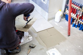 Pracownik rozprowadza klej na płytce podłogowej za pomocą pacy ząbkowanej. W tle widoczna instalacja grzewcza i biały pojemnik.