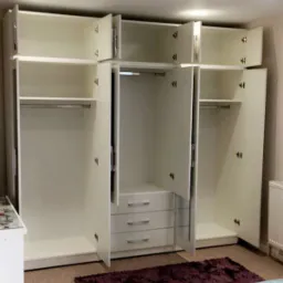 Biała szafa z otwartymi drzwiami w przedpokoju, widoczne p&oacute;łki, drążki i trzy szuflady, na podłodze mały, ciemny dywanik.