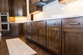 Ciemnobrązowe, drewniane fronty szafek kuchennych z prostymi, czarnymi uchwytami, jasny blat z szarymi żyłkami, parkiet w jodełkę, piekarnik w zabudowie po lewej stronie, wyciąg nad płytą grzewczą.