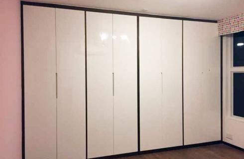 Szafa w przedpokoju z białymi, lakierowanymi frontami i ciemnobrązowymi obramowaniami, widoczna z boku, z fragmentem ściany w kolorze pudrowego różu i oknem z roletą w geometryczne wzory.