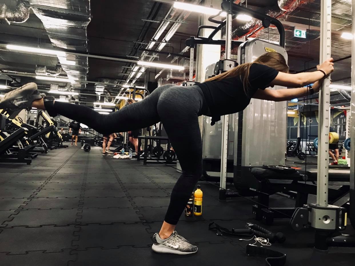 Kobieta w sportowym stroju wykonuje ćwiczenie odwodzenia nogi w tył na maszynie w siłowni, opierając się rękoma o pionową kolumnę maszyny.