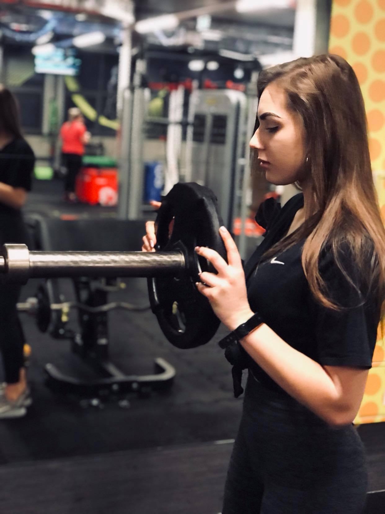 Kobieta z długimi włosami w czarnym stroju sportowym zakłada czarny obciążnik na sztangę w siłowni, widok z profilu.
