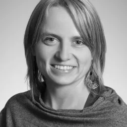 Portret uśmiechniętej kobiety z kolczykami o ażurowym wzorze, w szarym swetrze na jasnym tle.