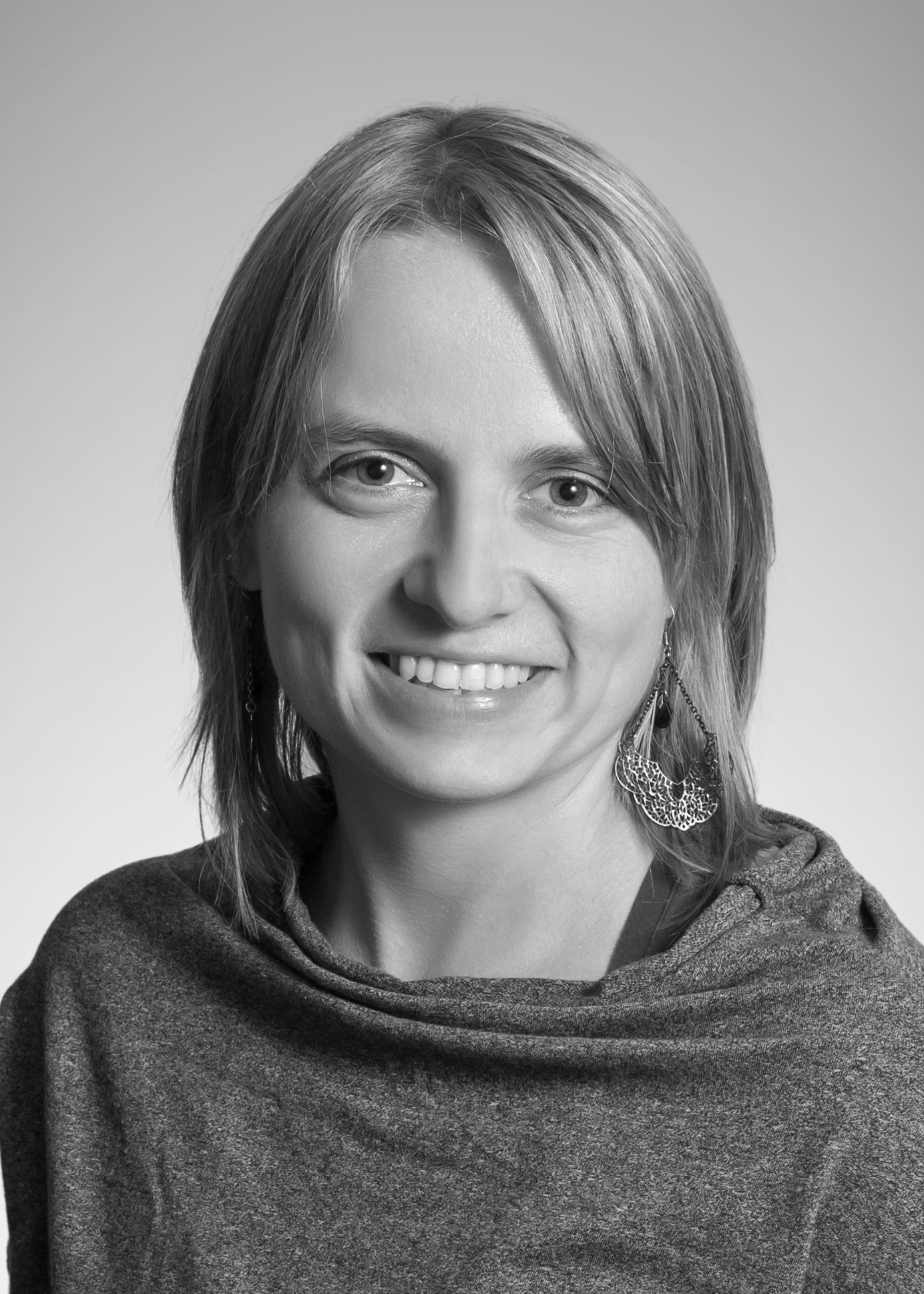 Portret uśmiechniętej kobiety z kolczykami o ażurowym wzorze, w szarym swetrze na jasnym tle.