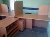 Biurka z przegrodami w jasnym odcieniu drewna, szafka z szufladami i regał otwarty, widoczne na tle szarej podłogi i zielonej ściany.