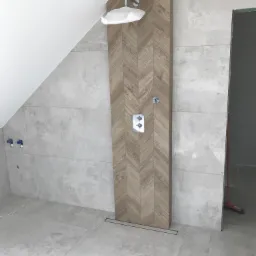 Nowoczesna kabina prysznicowa z panelami imitującymi drewno ułożonymi w jodełkę, szare płytki na ścianach i podłodze, widoczne zawory i deszczownica w folii ochronnej, sufit ze skosem.