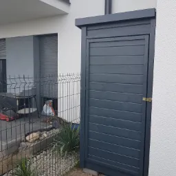 Drewniana szafa ogrodowa w odcieniach szarości, z poziomymi deskami i złotym zamkiem, przylegająca do białej ściany budynku, widoczna zza metalowego ogrodzenia.