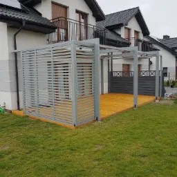 Nowoczesna konstrukcja tarasowa z szarego metalu z poziomymi panelami, przylegająca do domu z drewnianym tarasem, widok z trawnika.