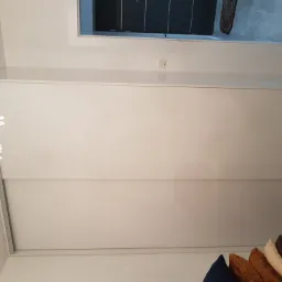 Biała szafa przesuwna z widocznymi kryształkami żyrandola w rogu kadru i fragmentem kolekcji poduszek dekoracyjnych w dolnej części zdjęcia.