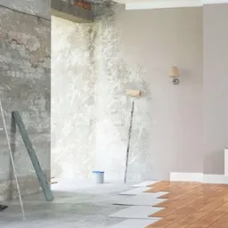 Remont pokoju: częściowo otynkowana ściana z cegłą, drabina, wałek do malowania, częściowo ułożony parkiet.