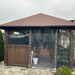 Drewniana altana z brązowym dachem krytym gontem bitumicznym i przezroczystymi ścianami foliowymi, wyposażona w ławkę i donicę z kwiatami, otoczona tujami i kostką brukową.