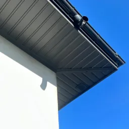 Szary okap dachu z zamontowanymi czarnymi rynnami i rurą spustową na tle błękitnego nieba, widok z dołu.