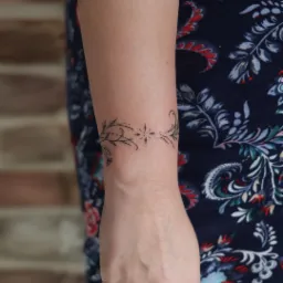 Delikatny tatuaż na nadgarstku kobiety, przedstawiający motyw roślinny z centralną gwiazdą, wykonany czarnym tuszem na tle wzorzystej, granatowej tkaniny.