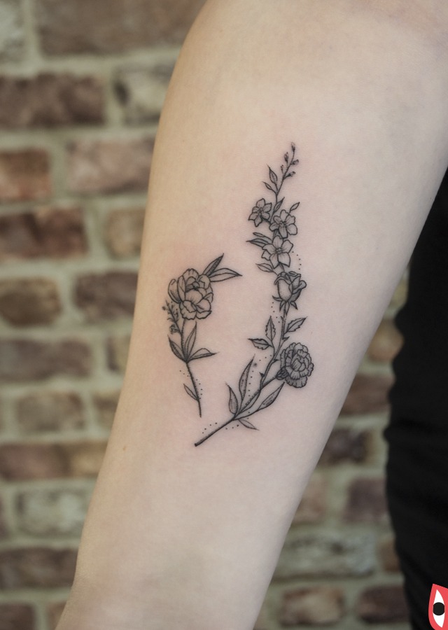Delikatny tatuaż na ramieniu przedstawiający dwie gałązki kwiatów w stylu minimalistycznym, z drobnymi detalami i czarnym tuszem, na tle ceglanej ściany.