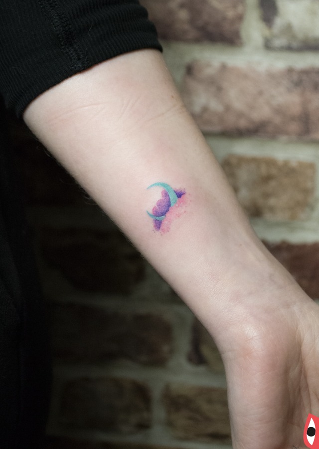 Delikatny, akwarelowy tatuaż na przedramieniu przedstawiający księżyc i chmurę w odcieniach fioletu i błękitu, wykonany w minimalistycznym stylu.