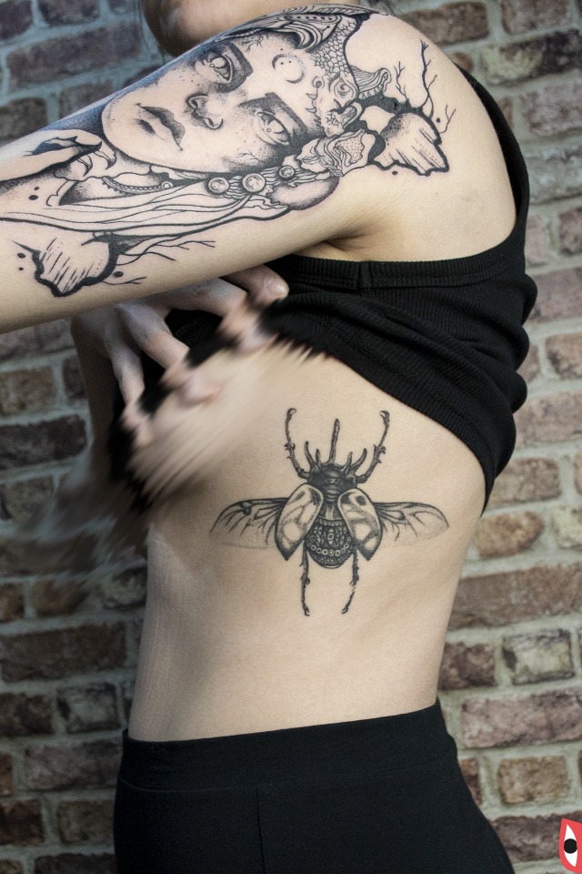 Kobieta z czarnym topem prezentuje dwa tatuaże: na ramieniu stylizowana twarz kobiety i na boku tułowia tatuaż chrząszcza skarabeusza na tle ściany z cegły.