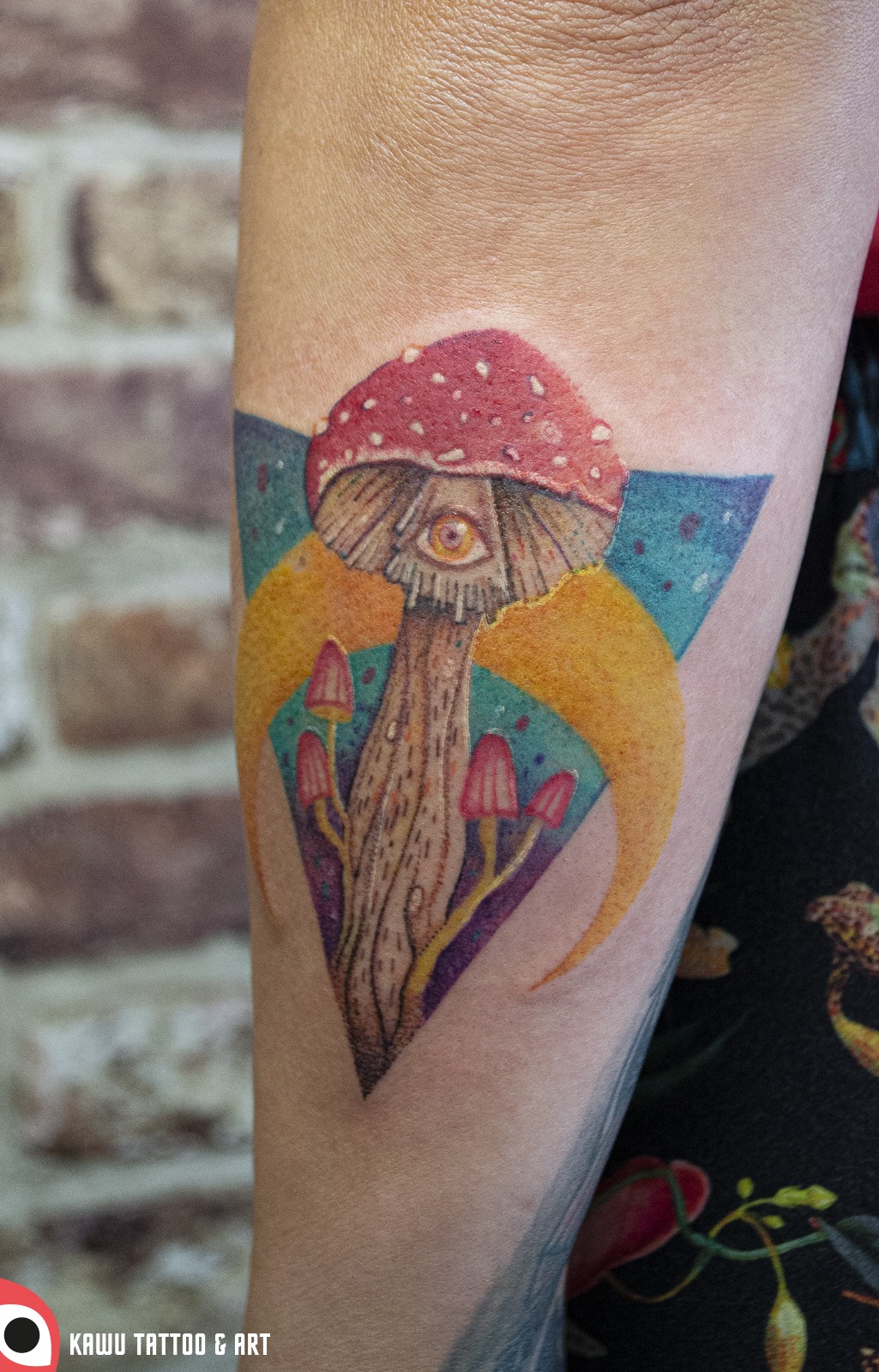 Kolorowy tatuaż na ramieniu przedstawiający muchomora z okiem w kapeluszu, umieszczony w geometrycznym tle z trójkątem i półksiężycami.