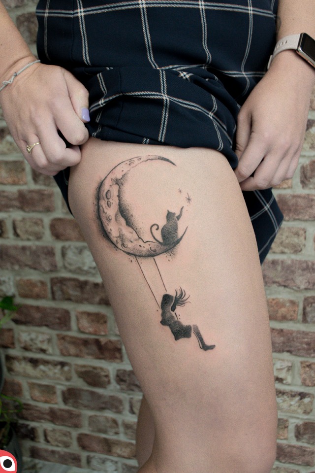 Czarno-biały tatuaż na udzie przedstawiający kota siedzącego na księżycu i dziewczynę na huśtawce, z ceglanym tłem.