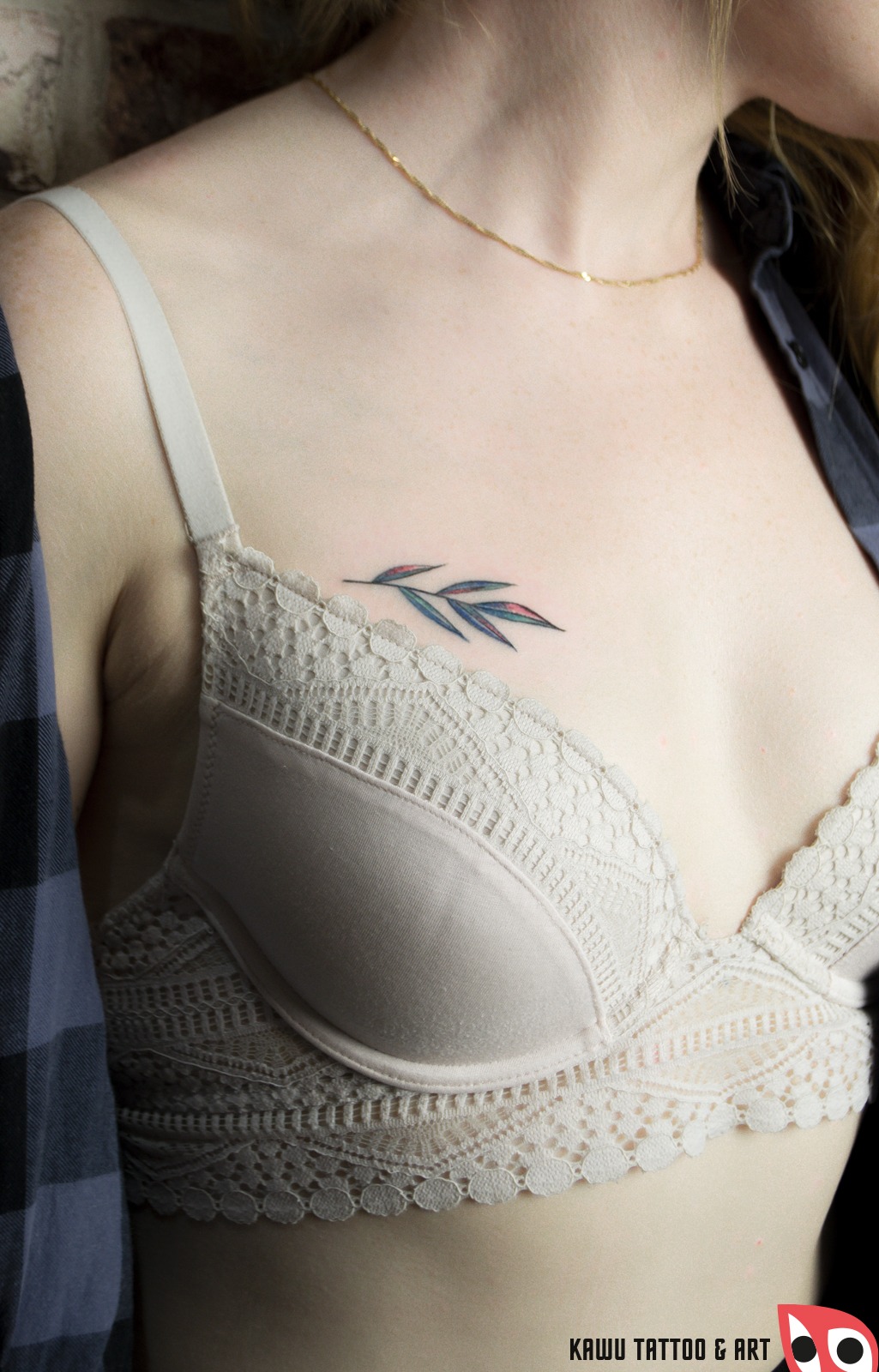 Delikatny tatuaż gałązki z liśćmi w kolorach niebieskim i różowym, umieszczony na dekolcie kobiety, widoczny fragment biustonosza z koronką i złoty łańcuszek.