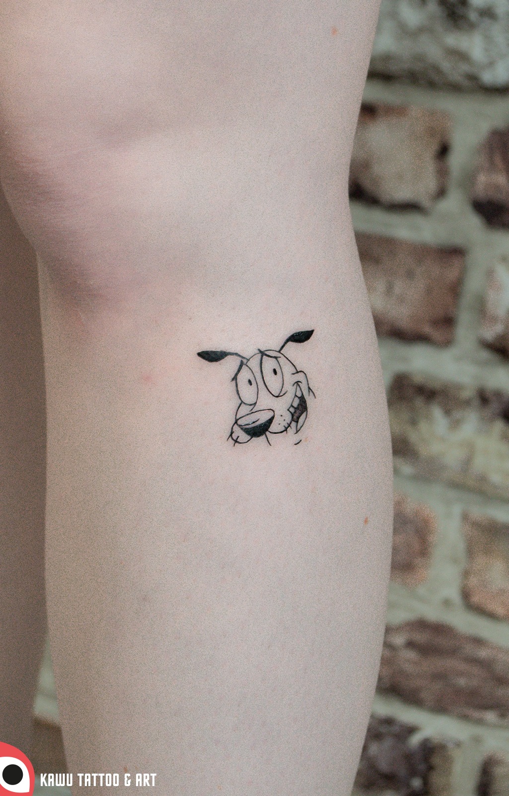 Czarno-biały tatuaż na nodze przedstawiający głowę psa z kreskówki, wykonany cienką linią, na tle ceglanej ściany.