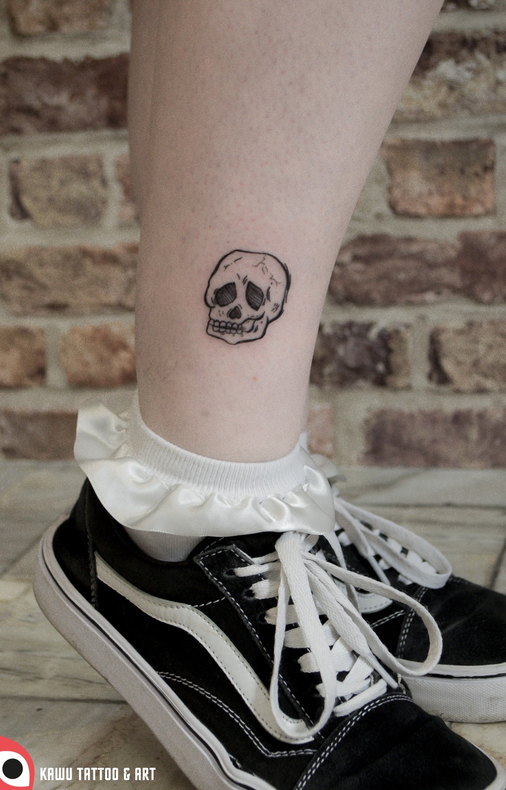 Drobny, czarny tatuaż przedstawiający czaszkę na nodze, tuż nad białą skarpetką z falbanką i czarno-białymi butami Vans, na tle ceglanej ściany.
