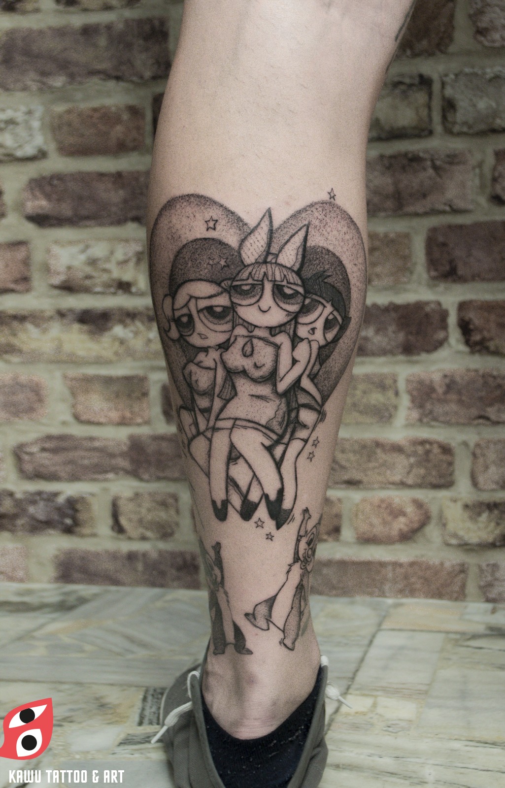 Tatuaż na łydce przedstawiający trzy postacie z kreskówki Atomówki, wykonany czarnym tuszem na tle serca z gwiazdami; widoczny fragment stopy w szarym trampku.