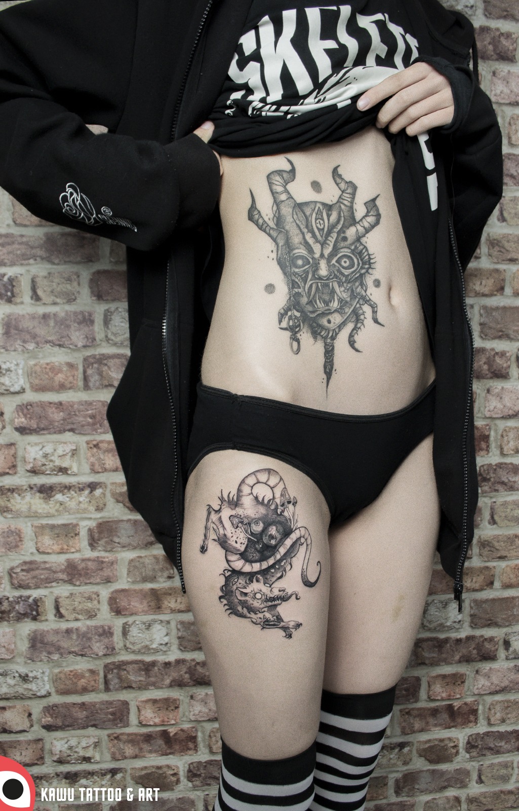 Dwie czarno-białe, szczegółowe grafiki tatuaży na ciele kobiety: demoniczna maska na brzuchu i fantastyczne stworzenie na udzie, na tle ceglanej ściany.