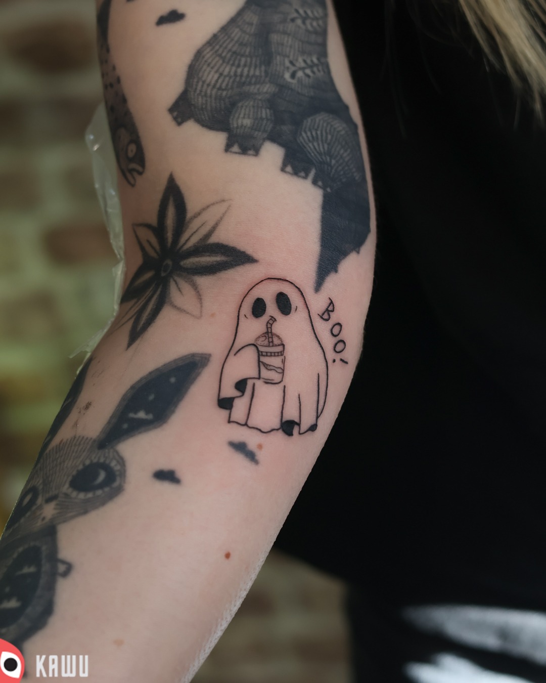 Tatuaż na ramieniu przedstawiający ducha trzymającego napój ze słomką i napisem 'Boo!', otoczony innymi tatuażami w stylu graficznym.
