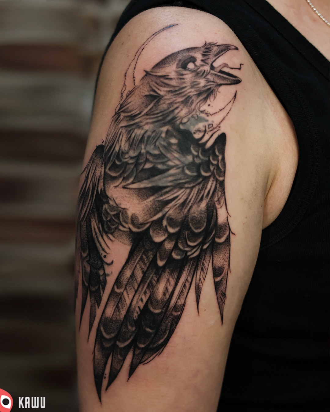 Tatuaż na ramieniu przedstawiający czarnego kruka z rozpostartymi skrzydłami, wykonany w technice dotwork z elementami graficznymi imitującymi księżyc w tle.