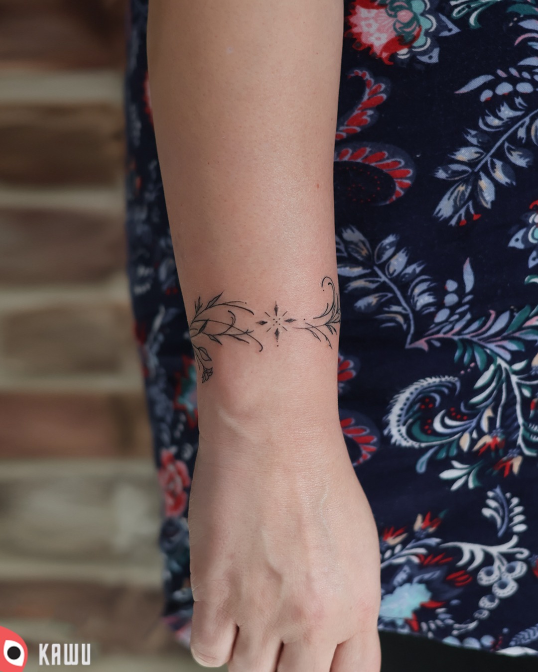 Delikatny tatuaż na nadgarstku kobiety, przedstawiający motyw roślinny z centralną gwiazdą, wykonany czarnym tuszem na tle wzorzystej, granatowej tkaniny.