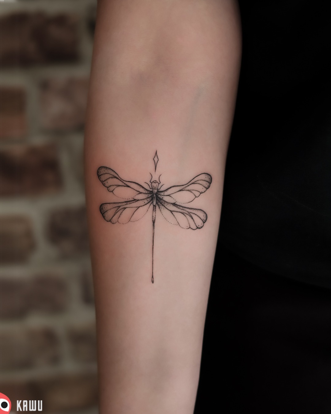 Delikatny tatuaż przedstawiający ważkę z geometrycznym akcentem na przedramieniu, wykonany czarnym tuszem na tle ceglanej ściany.