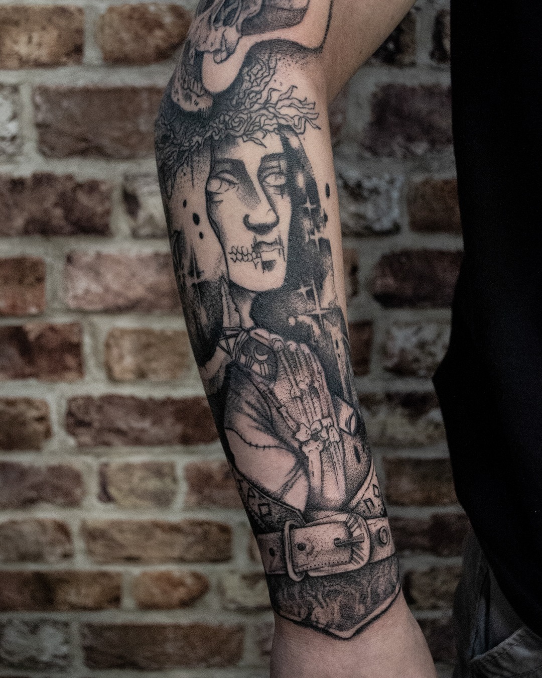 Tatuaż na ramieniu przedstawiający postać z koroną cierniową, złożonymi rękami i pasem, wykonany w stylu graficznym na tle ceglanej ściany.