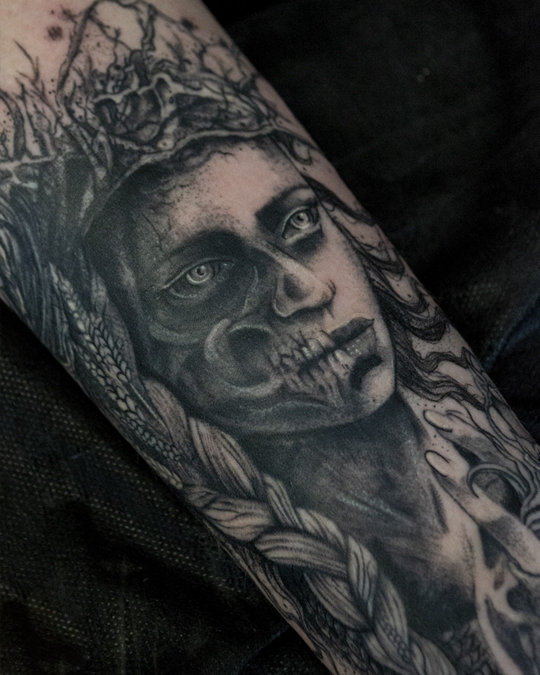 Tatuaż na ramieniu przedstawiający portret kobiety z elementami czaszki, wykonany w czerni i szarości, z detalami takimi jak warkocz i korona z motywami roślinnymi.